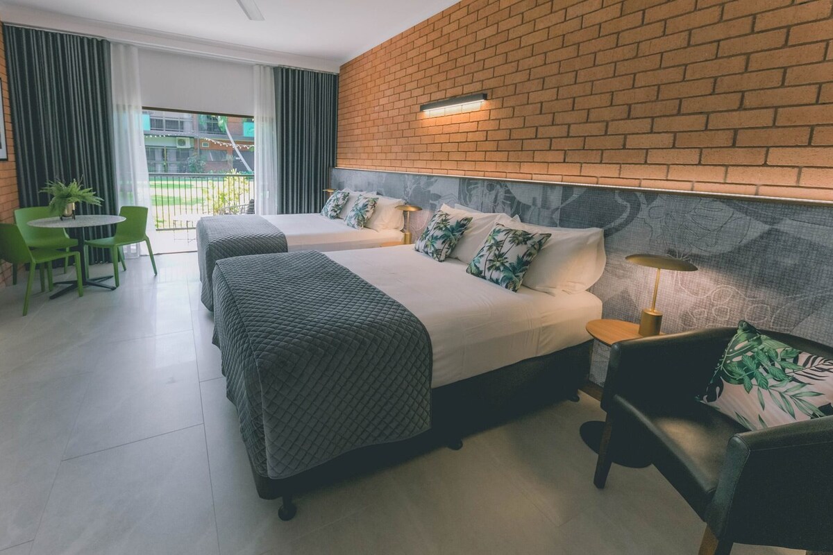 Habitación de hotel con pared de ladrillo decorativa, cama con cubrecama acolchado, escritorio con silla y balcón con vistas.