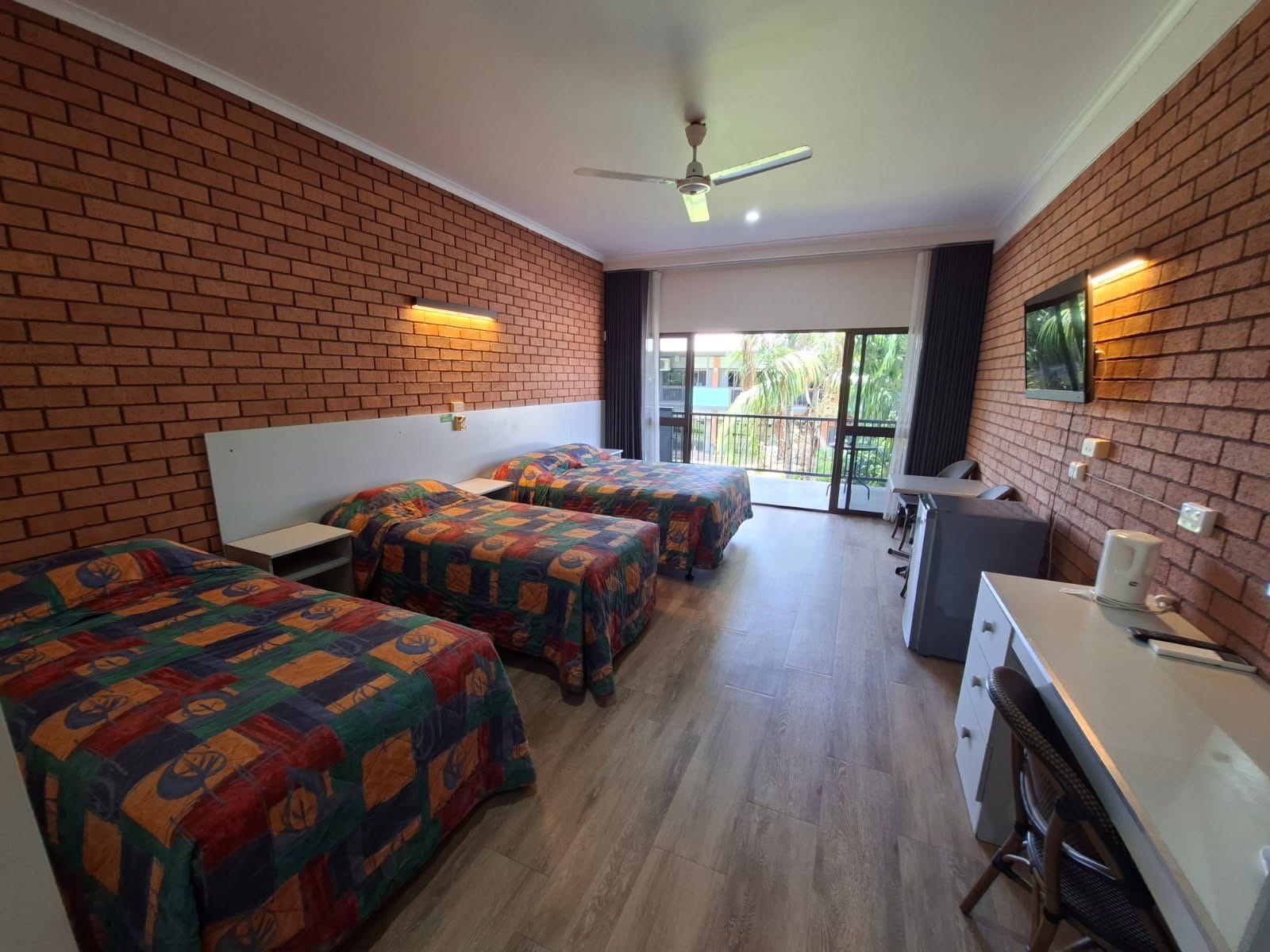 A hotel room with three beds, a desk, a chair, a ceiling fan, and a view of the outdoors.