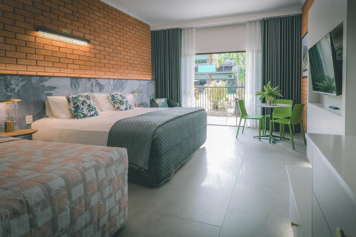 A hotel room with a brick wall, a bed with a quilted cover, a second bed, a green chair, a small table, and a TV on the wall.
