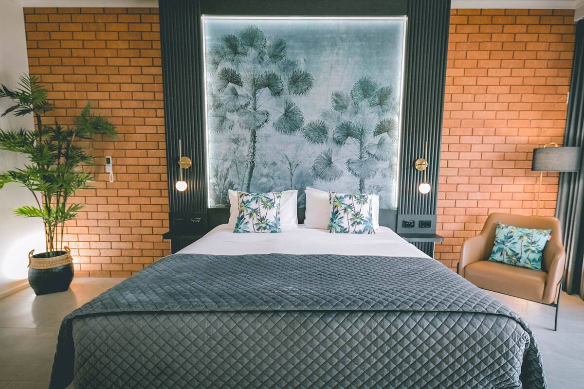 A modern bedroom with a large bed, a potted plant, a sofa, and a large window with a view of palm trees.