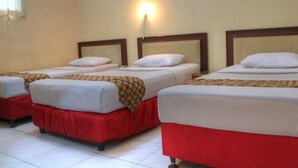 Quarto de hotel com três camas de solteiro, cada uma com saia vermelha e roupa de cama branca. Uma luminária fixada na parede ilumina o espaço.