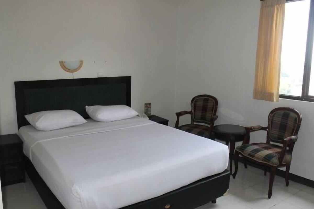A neatly made bed with white linens, a dark headboard, and a small bedside table with a lamp. There is a plaid-patterned chair and a small round table with a chair next to the bed.