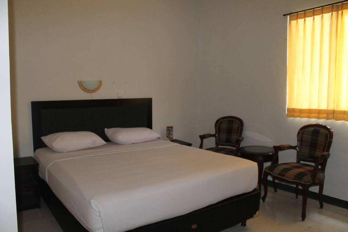 A neatly made bed with a headboard, a chair, a small table, and a window with curtains.