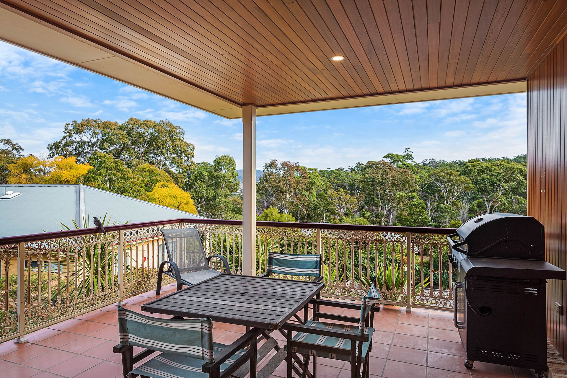 An outdoor patio with a table and chairs, a grill, and a view of trees and a building in the background.