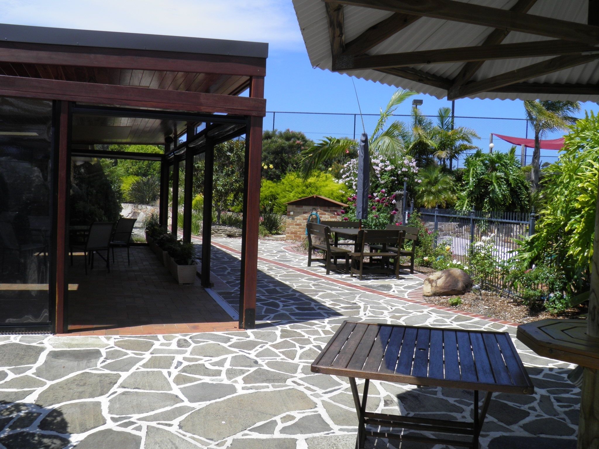 A covered patio area with stone flooring, outdoor seating, and a view of a garden with various plants and a fence.