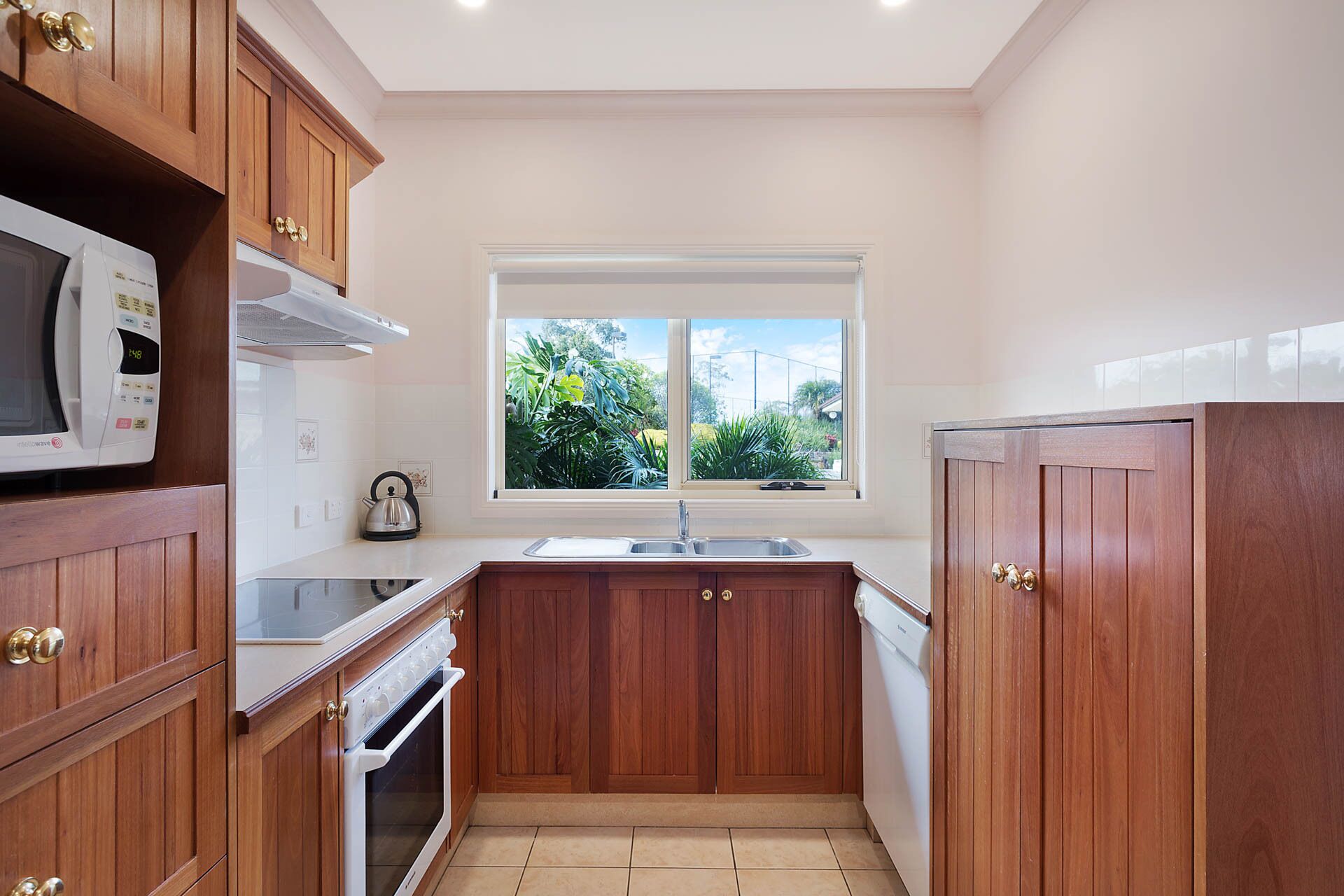 A compact kitchen with wooden cabinets, a microwave, an oven, a sink, and a window with a view of greenery.