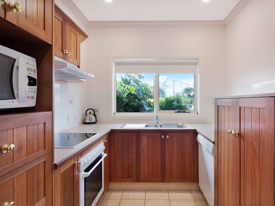A compact kitchen with wooden cabinets, a microwave, an oven, a sink, and a window with a view of greenery.