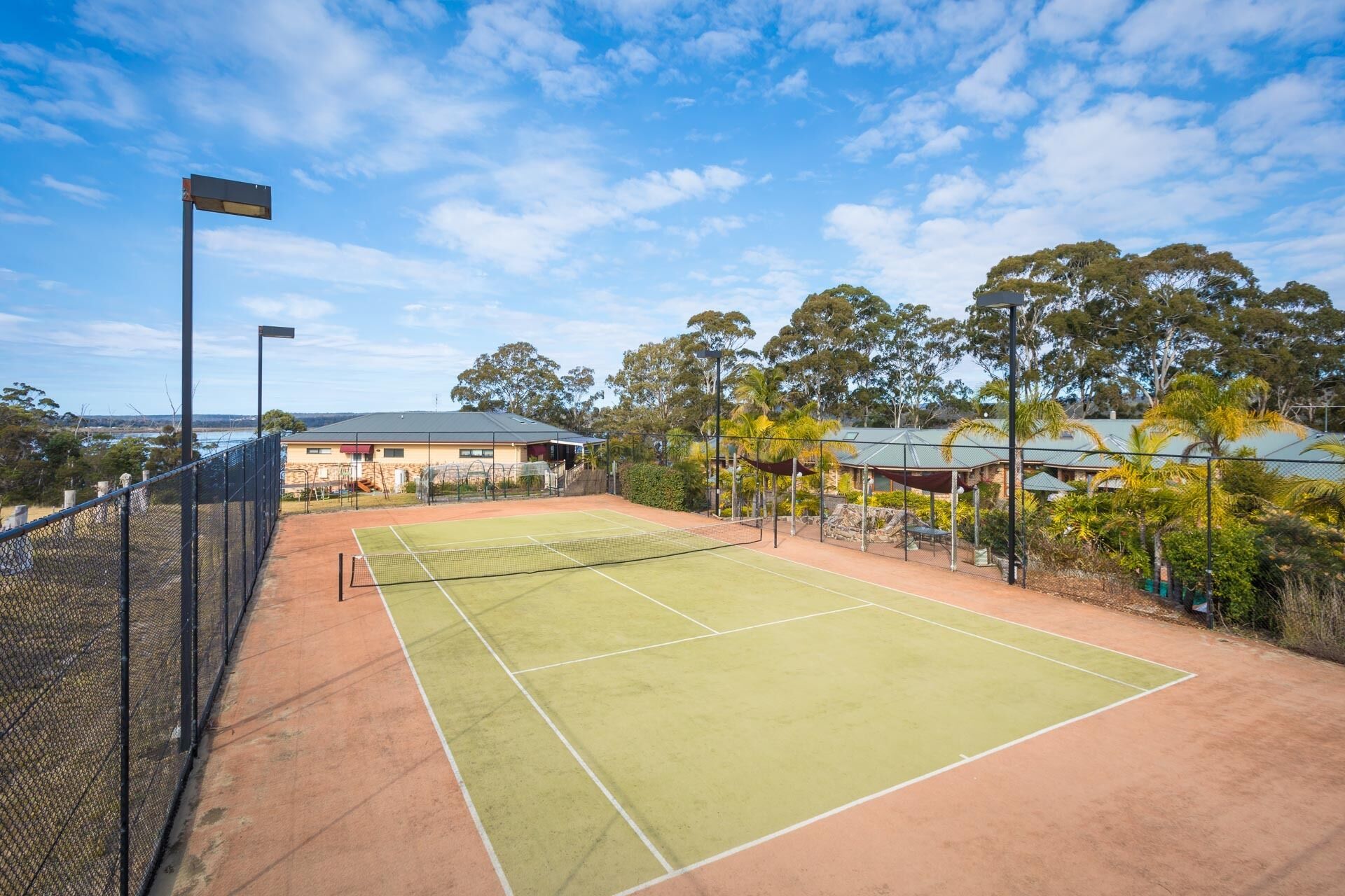 An outdoor tennis court with a net, surrounded by a fence and trees.
