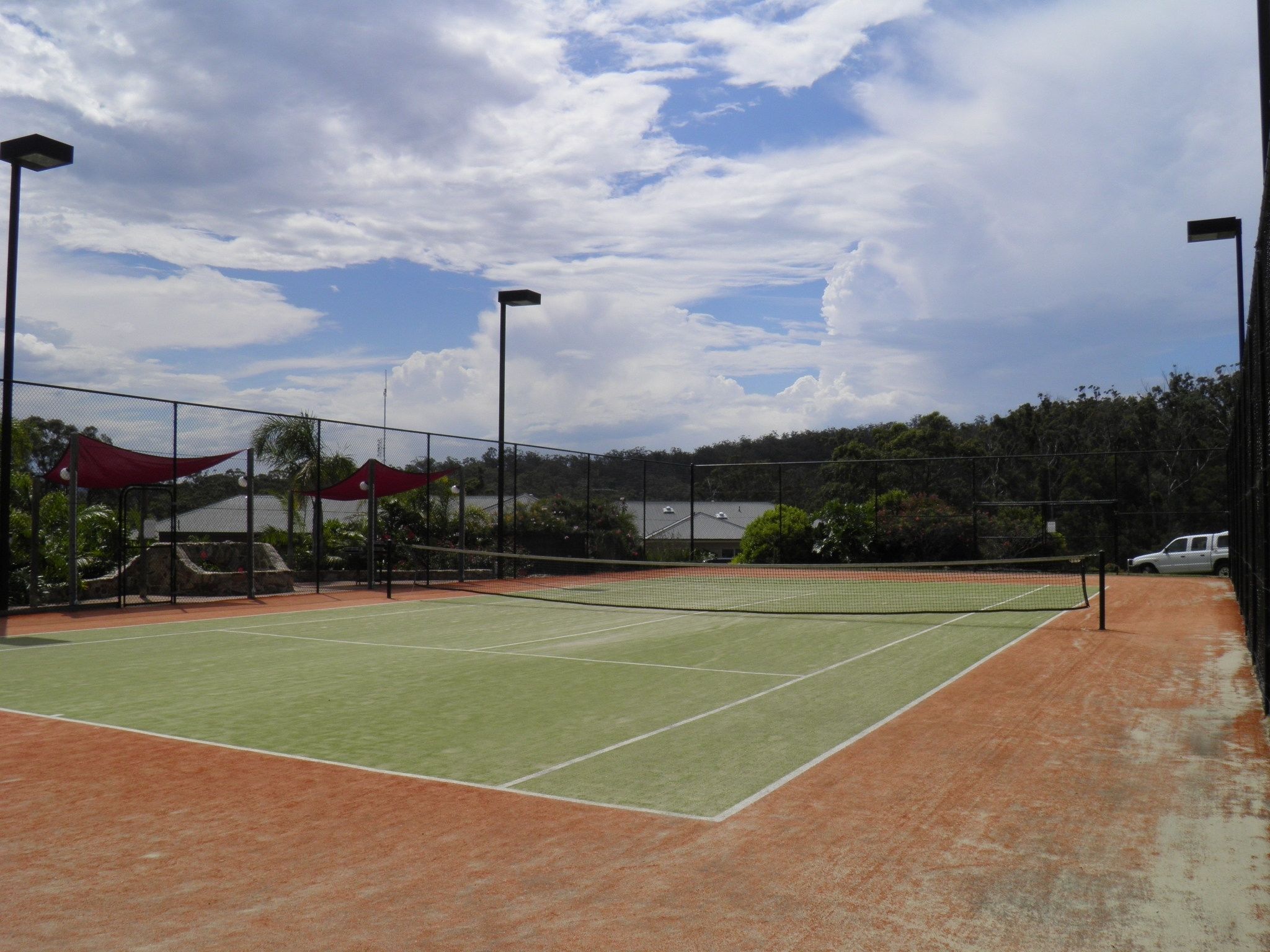 An outdoor tennis court with a net, surrounded by a fence and shaded by red canopies.