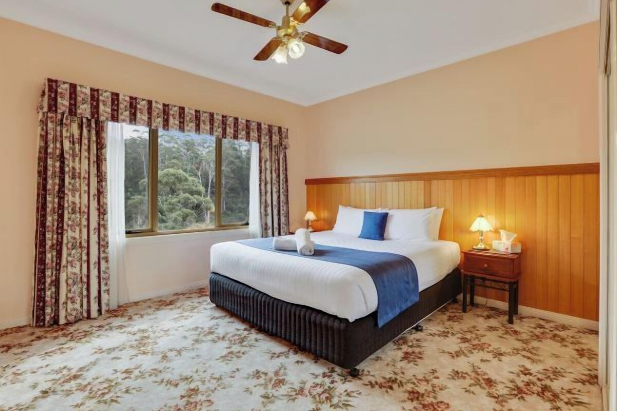 Un dormitorio con cama, mesita de noche, ventilador de techo y ventana con cortinas.