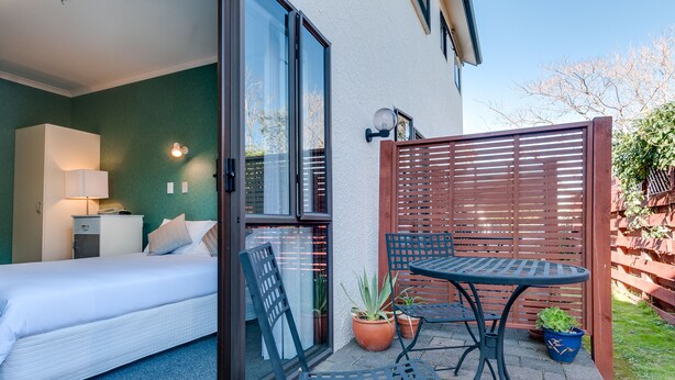 Ein Schlafzimmer mit Bett, Nachttisch, Lampe und Blick durch eine Schiebetür auf eine Außenpatio-Terrasse mit Tisch, Stühlen und Topfpflanzen.