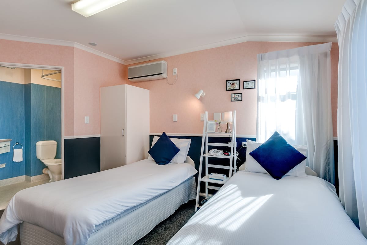 A hotel room with two single beds, a wardrobe, a desk with a chair, a small shelf, and a window with curtains.