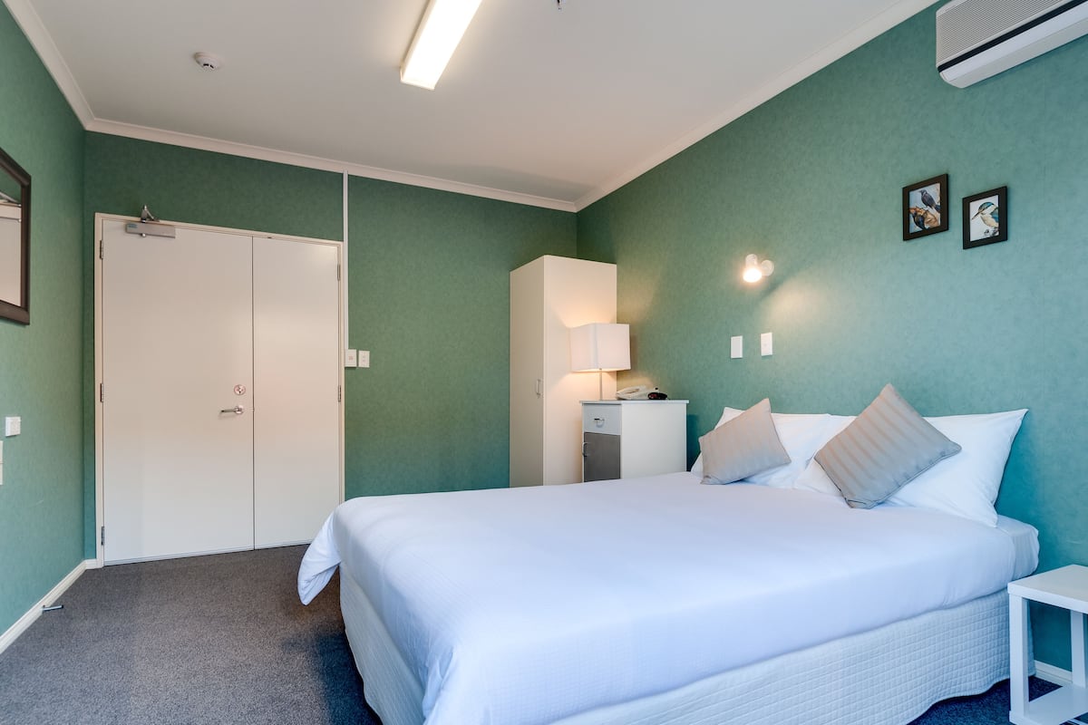 A hotel room with a large bed, a bedside table, a wardrobe, a desk with a lamp, and wall-mounted artwork.