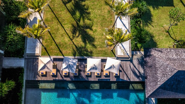 An aerial view of a pool area with lounge chairs and umbrellas, surrounded by palm trees and a wooden deck.