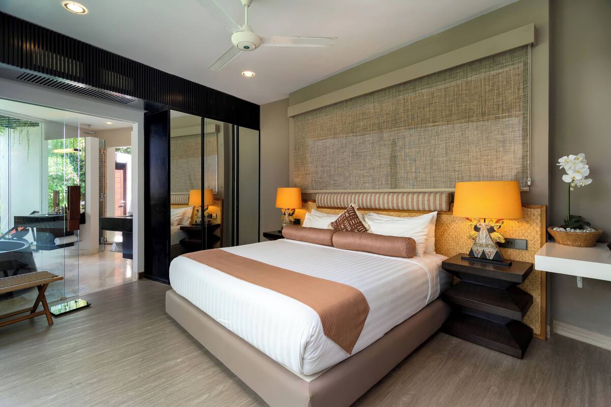 A modern hotel room with a large bed, two bedside lamps, a ceiling fan, and a view of the outdoors.
