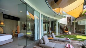 A modern outdoor living area with a curved glass wall, a wooden deck, and a seating arrangement including a sofa and a chair. - Villa Toraja by Lifestyle Retreats (Canggu)