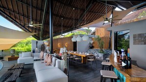 A spacious outdoor living area with a wooden deck, white sofas, a dining table, and a bar area with stools. - Villa Toraja by Lifestyle Retreats (Canggu)