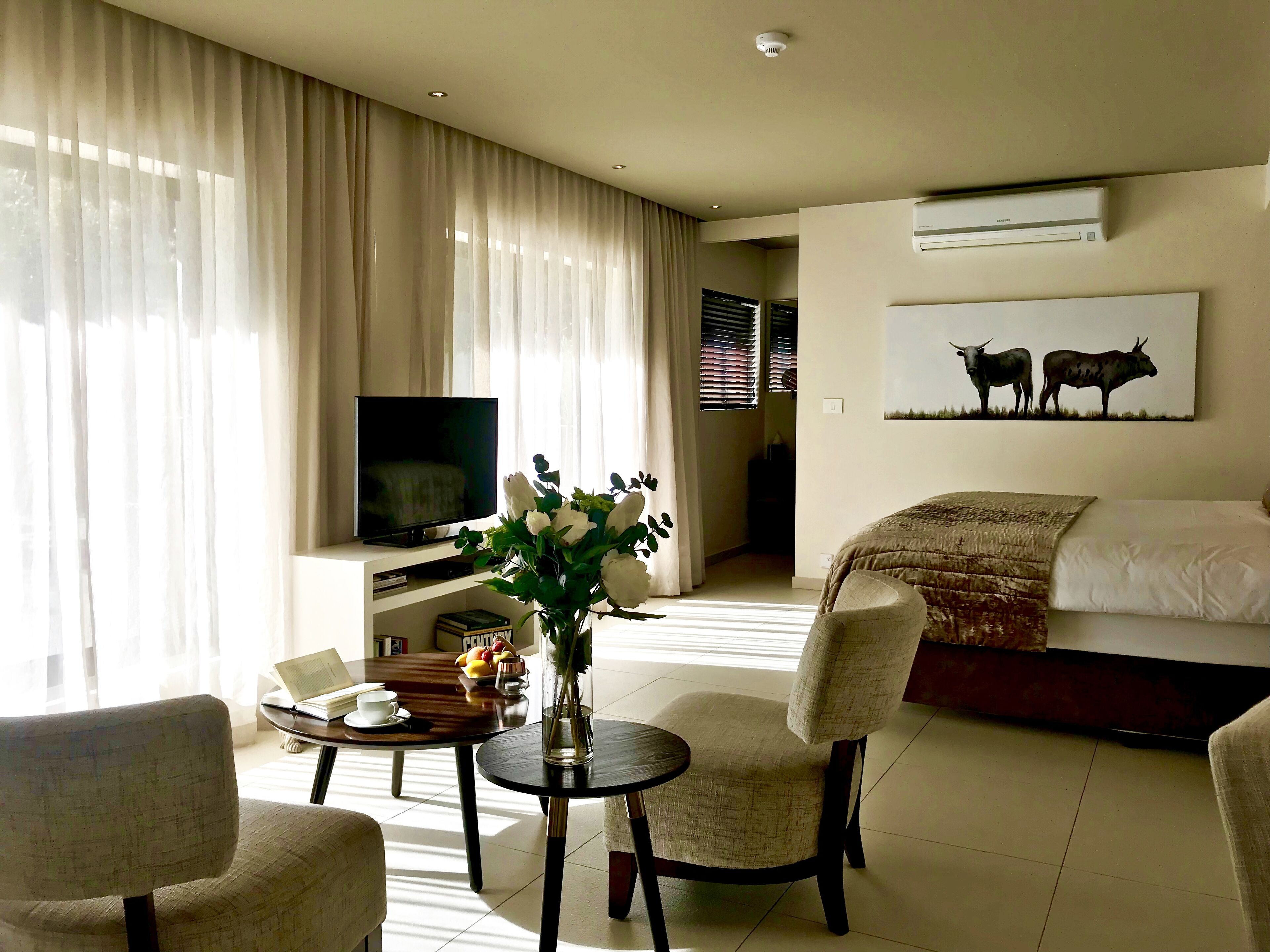 A hotel room with a TV, a dining table with chairs, a sofa, and a painting on the wall.