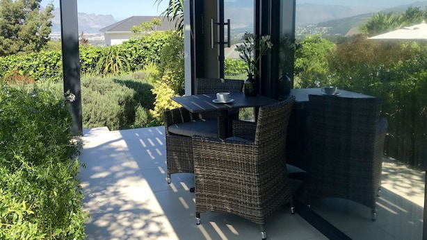 A balcony with wicker furniture, a table, and chairs, overlooking a lush garden and distant mountains. - Cape Vermeer (Cape Town)