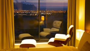 A hotel room with a large window offering a city view at dusk. Inside, there is a bed with a white comforter, a bedside table with a lamp, and two chairs. - Cape Vermeer (Cape Town)