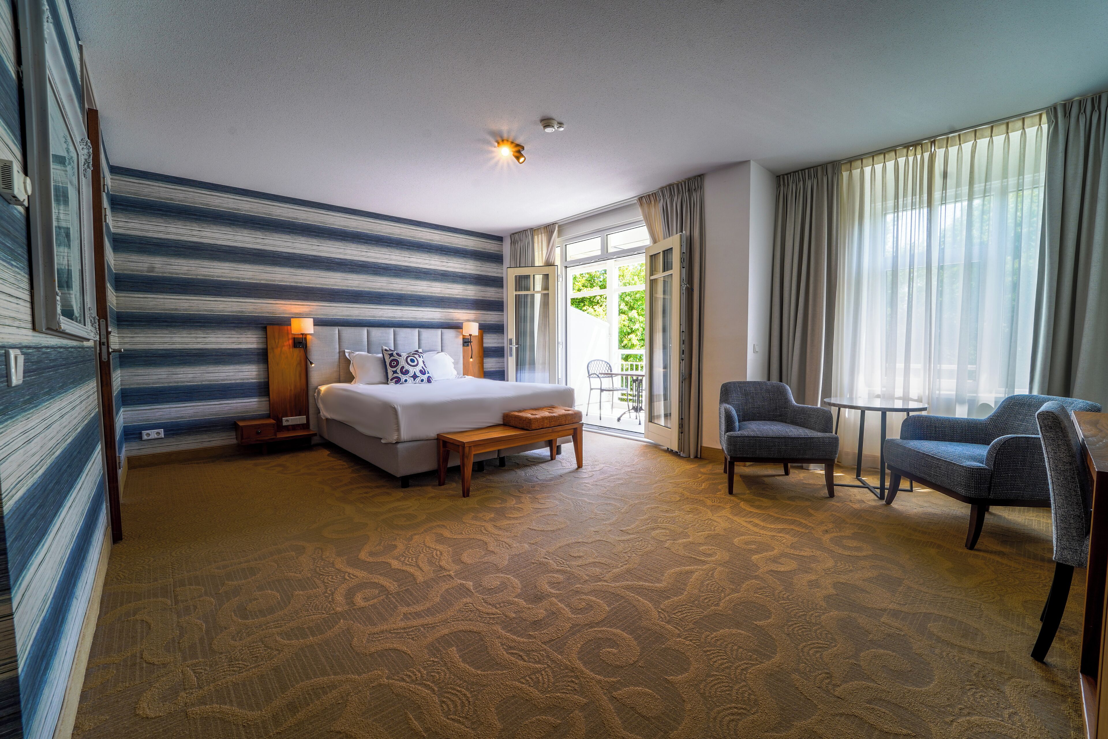 Une chambre d’hôtel avec un lit, deux fauteuils, une petite table et une grande fenêtre avec des rideaux.