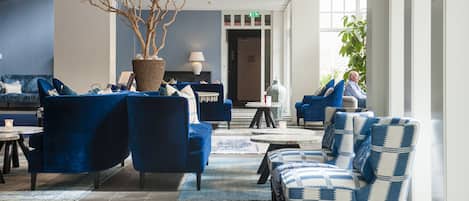 A modern lobby with blue sofas, a large potted tree, and a staircase leading to an upper floor.