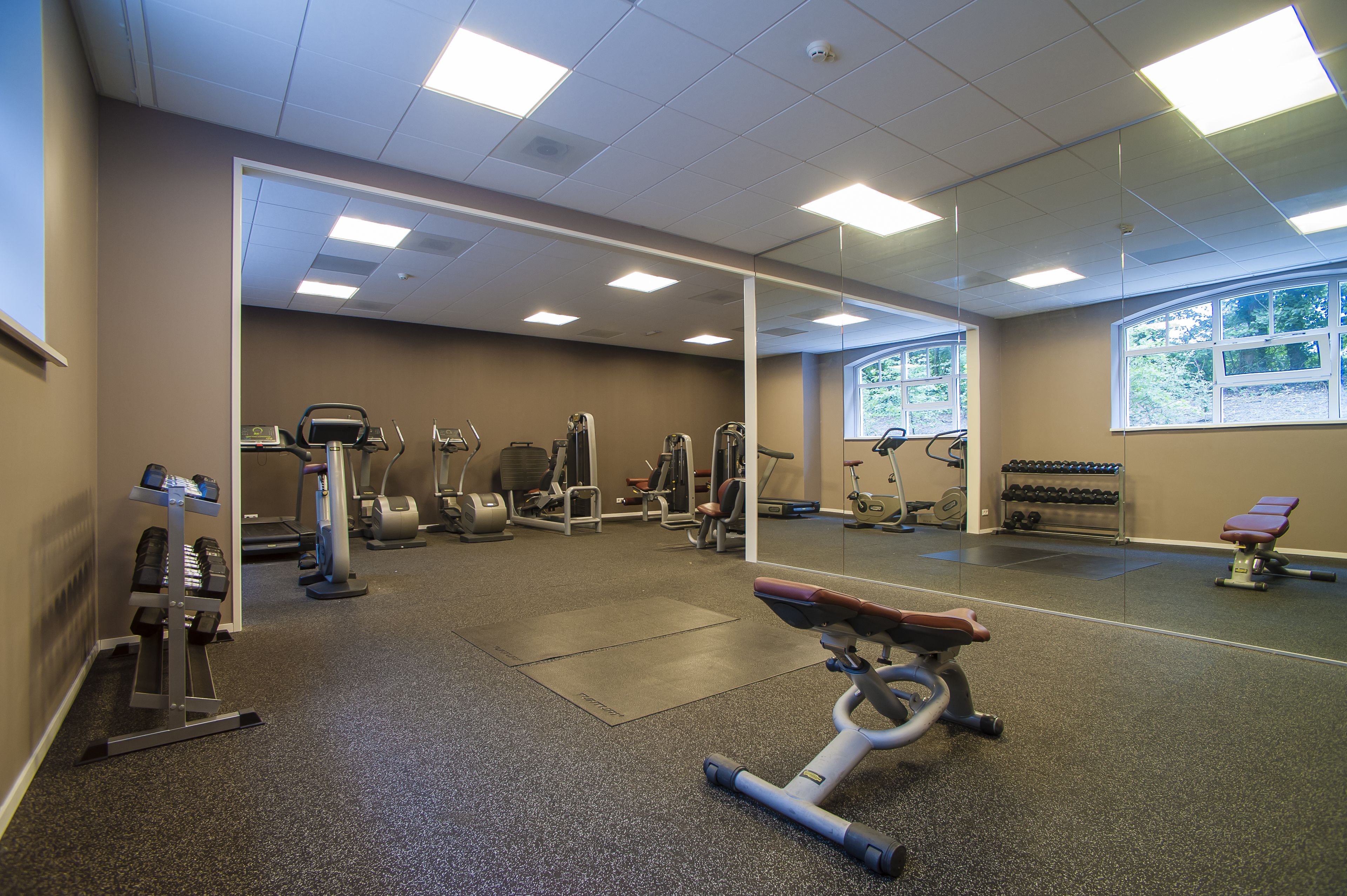 A gym with various exercise equipment, a mirrored wall, and a window with a view of trees.