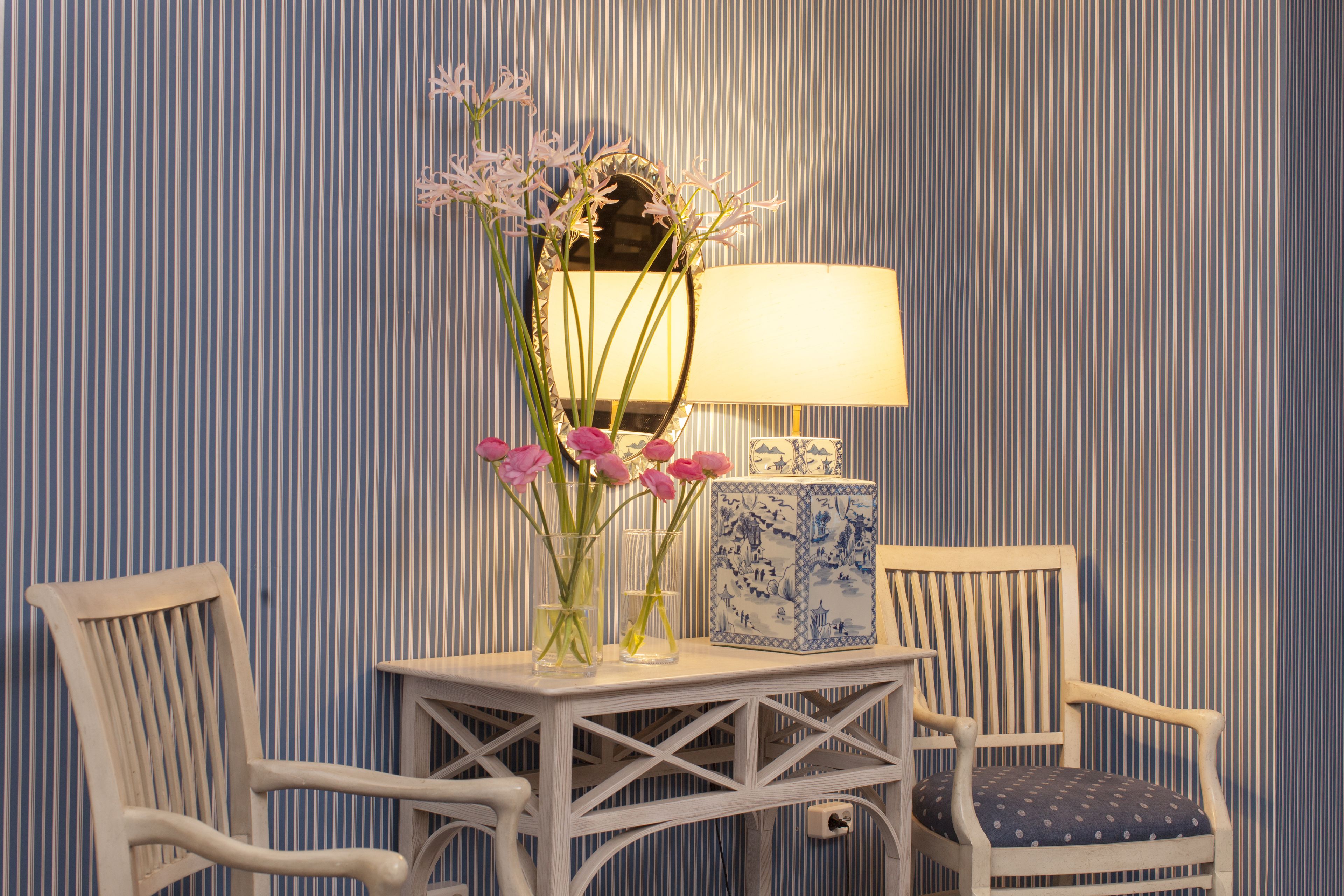 A room with striped wallpaper, a white table with a mirror, a vase with flowers, a lamp, and two chairs.
