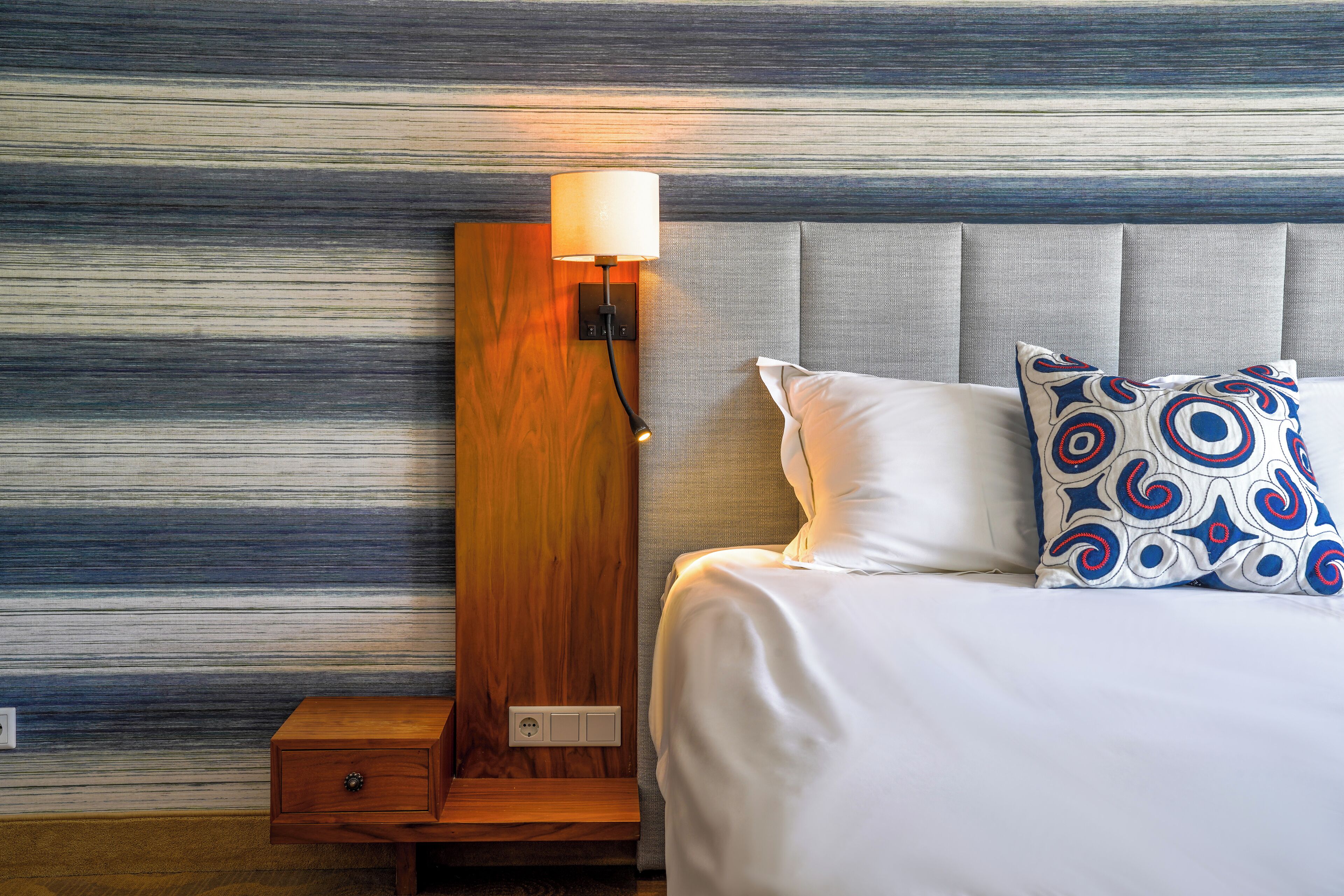 A neatly made bed with white bedding and patterned pillows, a bedside table with a lamp, and a wall with horizontal stripes.