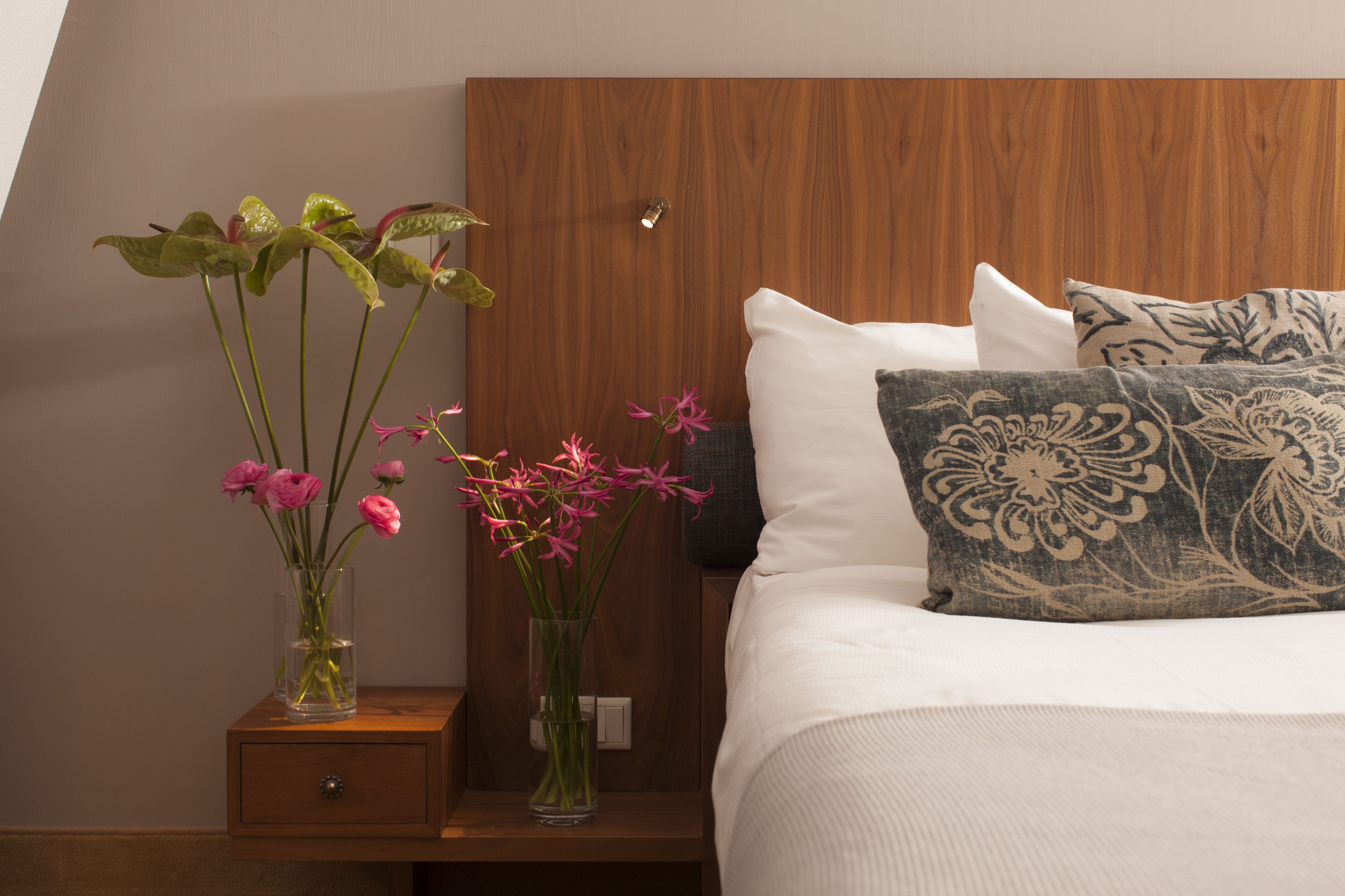 A neatly made bed with patterned pillows, a wooden headboard, and two vases with flowers on a bedside table.