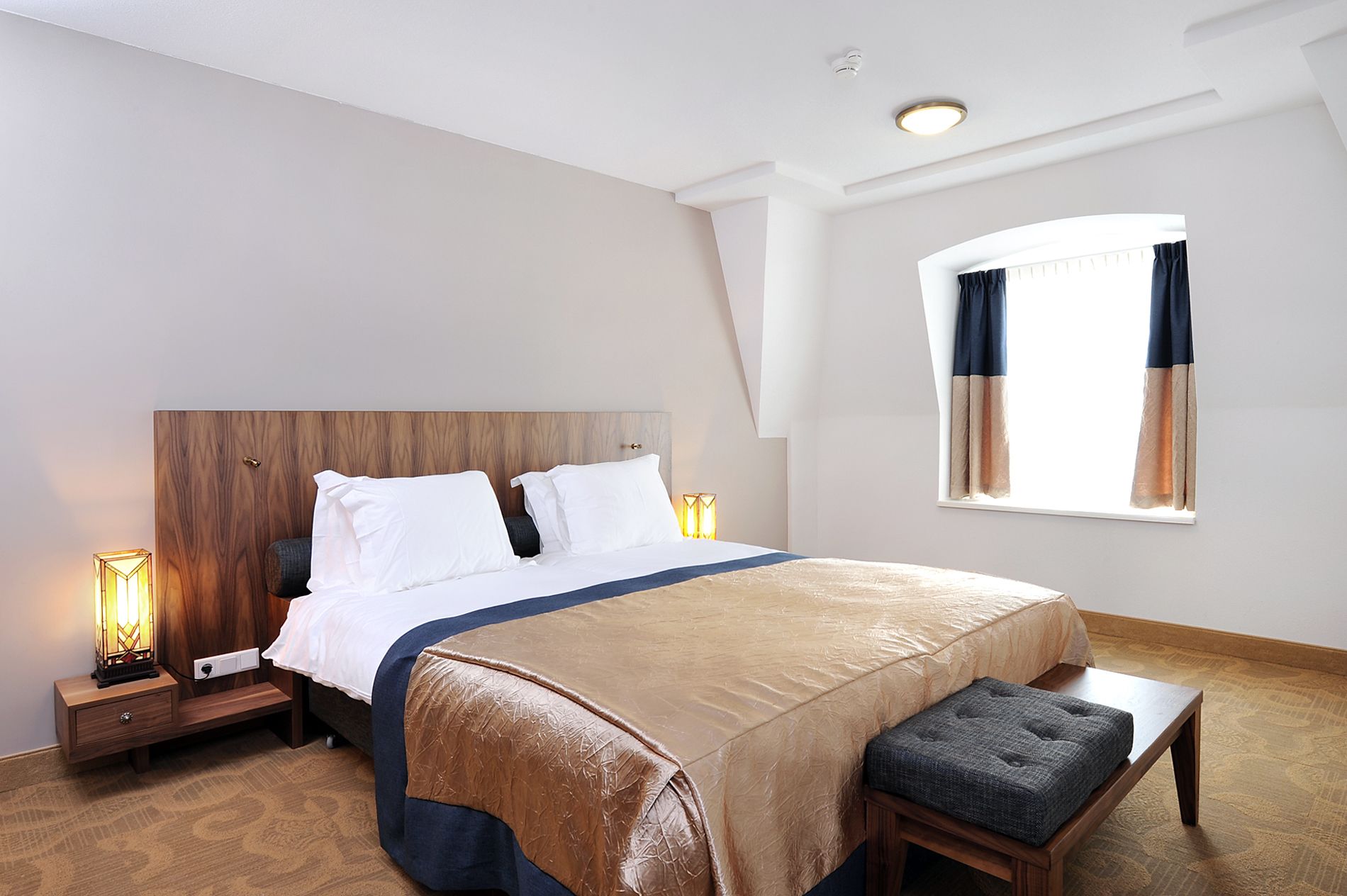 Moderni hotellihuone, jossa on suuri sänky, puinen pääty, yöpöytävalaisimet, penkki ja ikkuna, jossa on verhot.