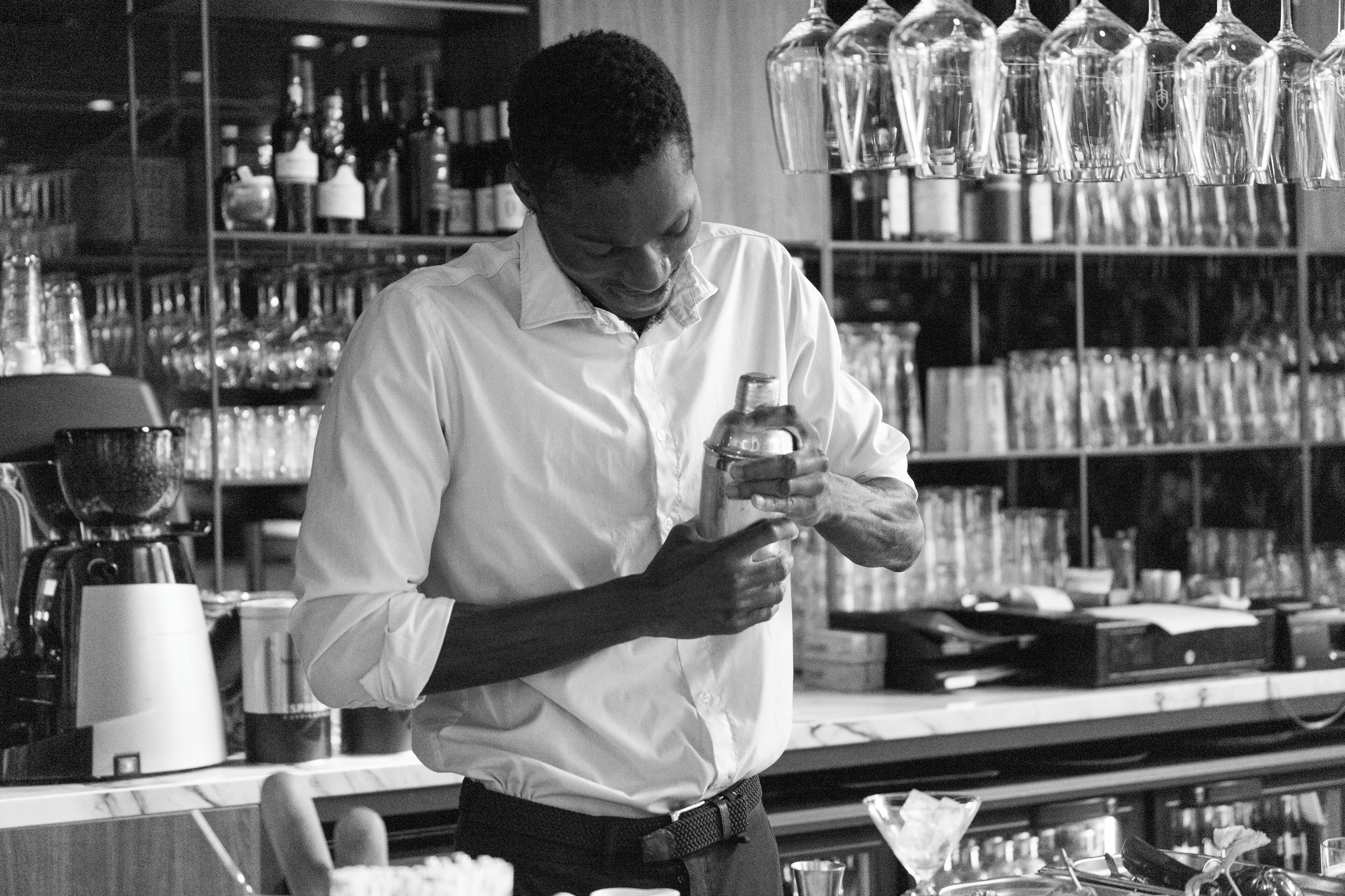 A person in a white shirt preparing a drink at a bar with shelves of bottles and glasses in the background.