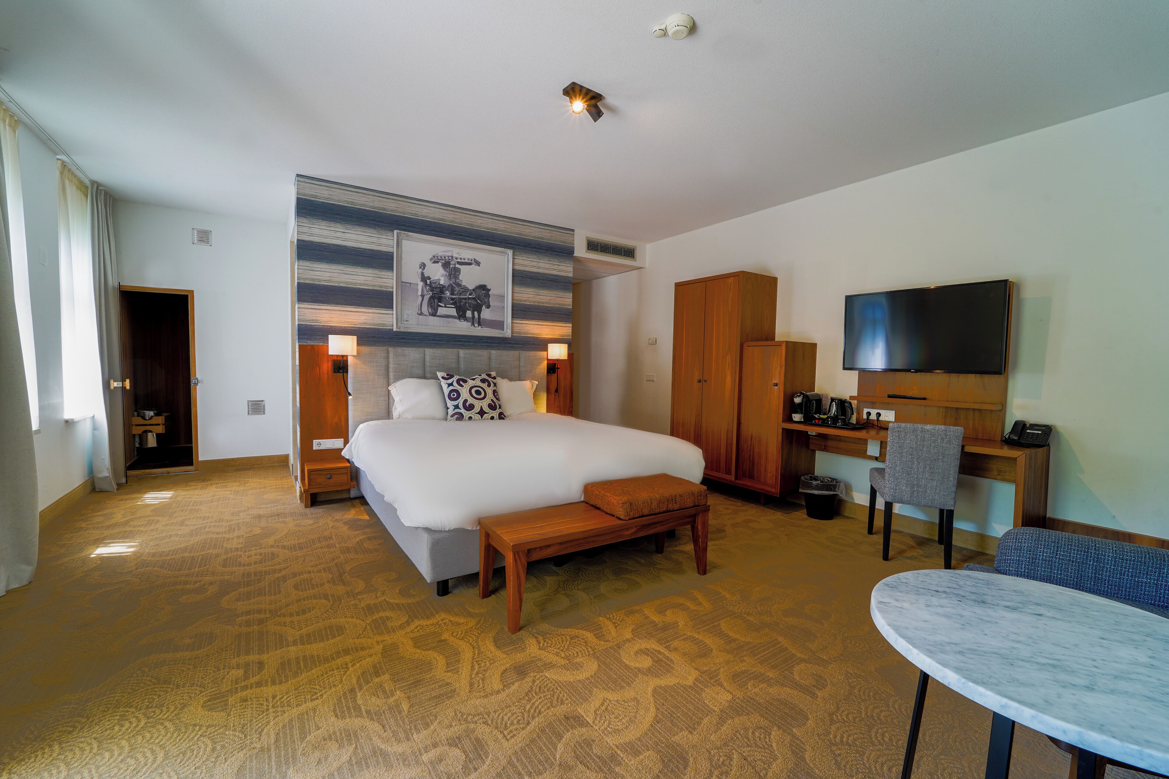 Hotelkamer met een groot bed, een bureau met stoel, een televisie en een kamerbreed tapijt met een patroon.