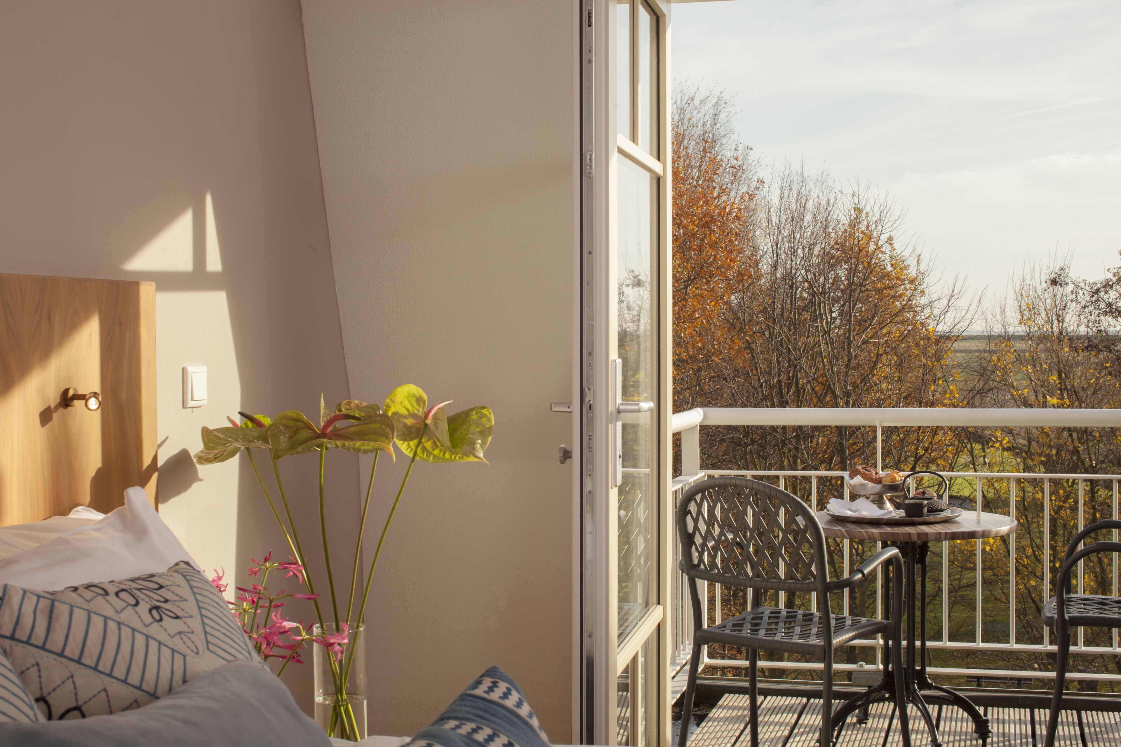 A balcony with a table and chairs, a bed with patterned pillows, and a view of trees.