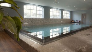 Uma piscina interior ampla com chão em mosaico, grandes janelas e uma planta em primeiro plano.