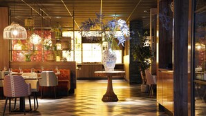 Um restaurante com chão xadrez, cadeiras listradas, uma mesa central com um vaso e luminárias pendentes.