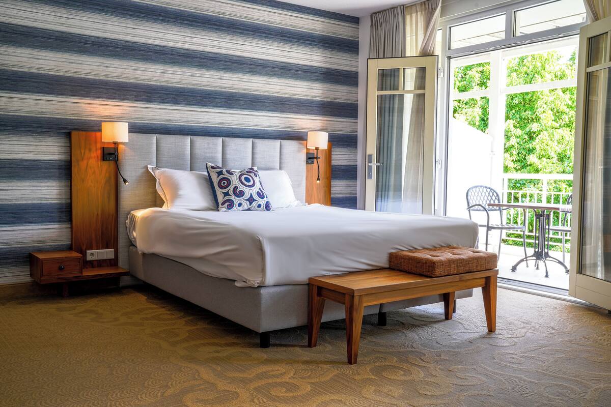 Ein Schlafzimmer mit einem großen Bett, zwei Nachttischen, einer Bank und Blick ins Freie.