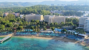An aerial view of a coastal resort with multiple buildings, a beach with sun loungers, and a pier extending into the water. - Özkaymak Select Hotel - Ultra All Inclusive  (Alanya)