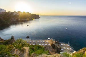 A coastal hotel with a sunlit terrace overlooking the sea, featuring sun loungers and a view of the coastline.