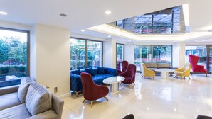 Un hall moderno con un sofá seccional azul, sillas rojas y amarillas, una mesa redonda y ventanas amplias que ofrecen vistas a zonas verdes.