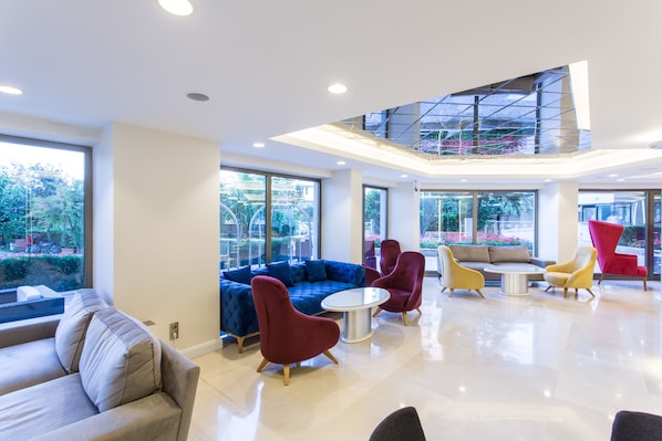 Un hall moderno con un sofá seccional azul, sillas rojas y amarillas, una mesa redonda y ventanas amplias que ofrecen vistas a zonas verdes.