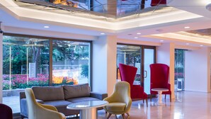 A modern lobby with a sofa, armchairs, and a small table. Large windows offer views of greenery outside. - Falcon Hotel (Antalya)