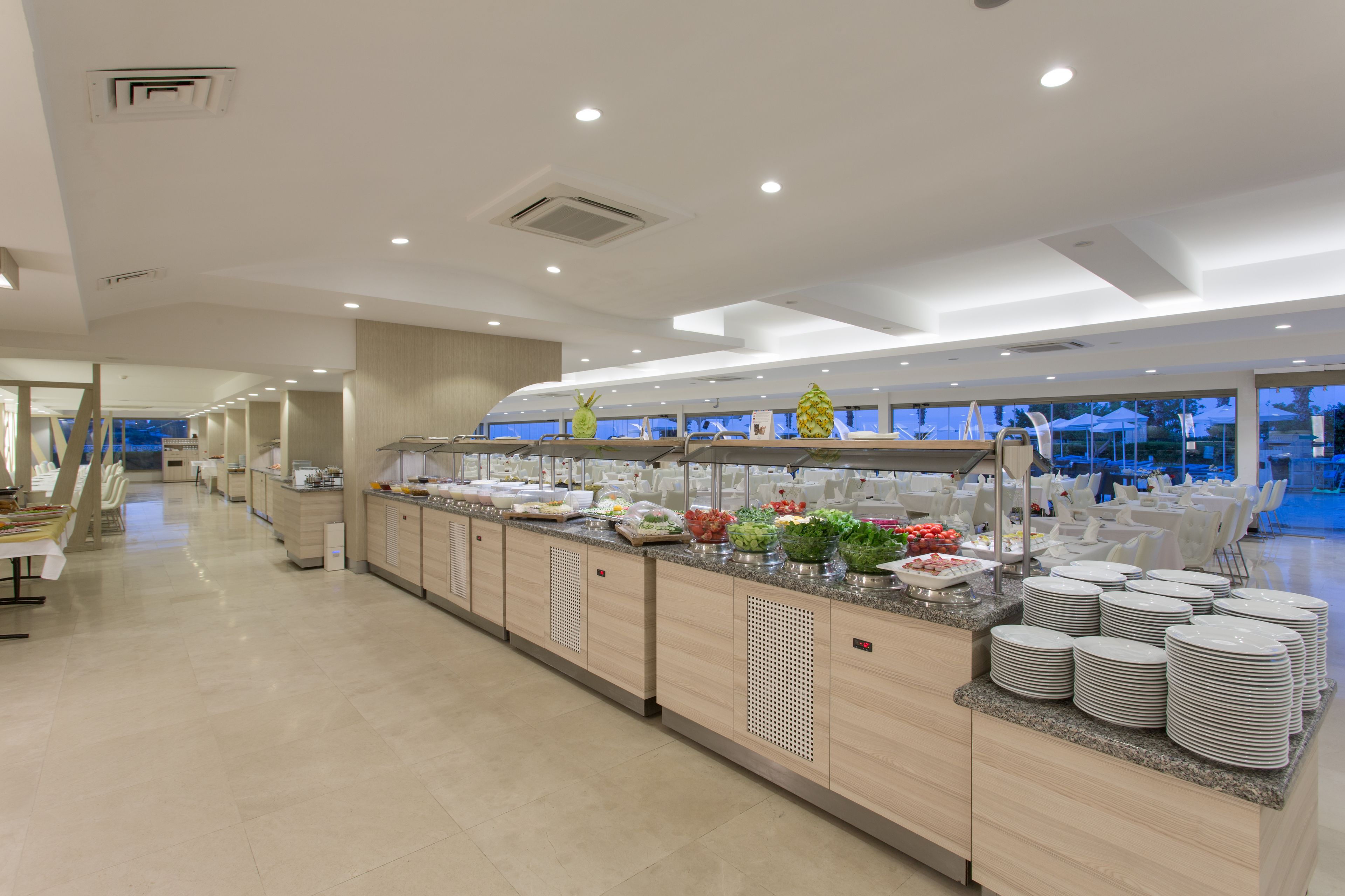 Un buffet proposant une variété de plats et de piles d’assiettes, avec un aménagement intérieur moderne comprenant un long comptoir et un espace salon.