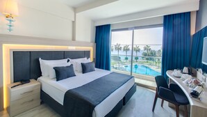 A hotel room with a bed, bedside table, desk, and a view of a pool and palm trees. - Falcon Hotel (Antalya)