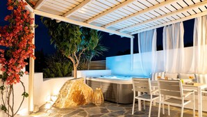 Un patio al aire libre con jacuzzi, suelo de piedra, mobiliario blanco y una pérgola con telas colgantes.