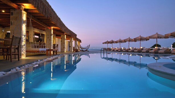 Zona de piscina con sillones y sombrillas, una estructura con techo de paja y vistas al mar.