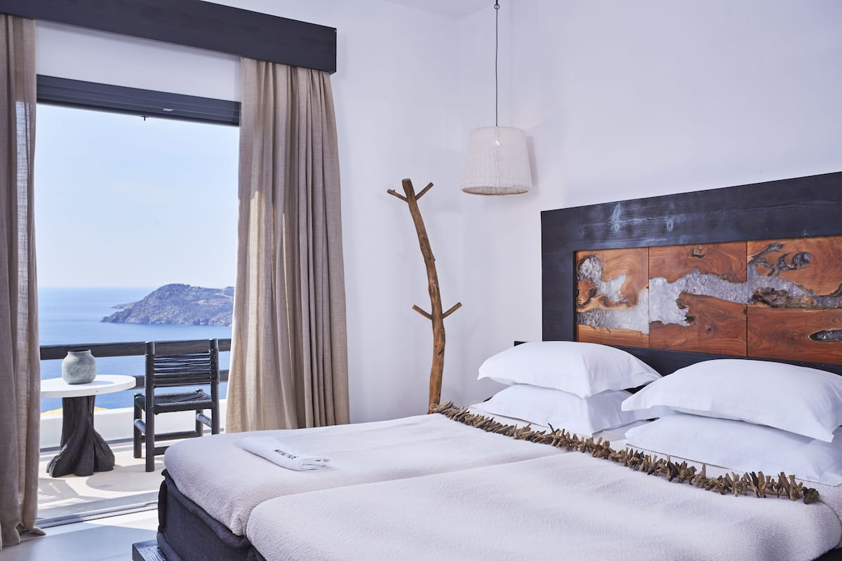 A hotel room with a large bed, a wooden headboard, a view of the sea, and a balcony with a table and chair.