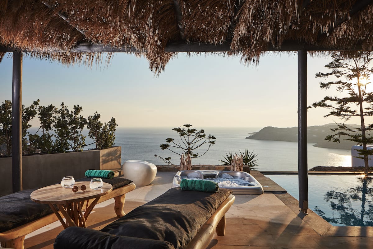 A sunlit outdoor seating area with a view of the sea, a thatched roof, and a pool.