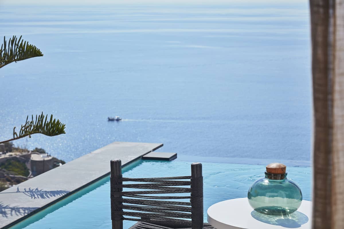 An infinity pool with a view of the ocean, a wicker chair, and a glass bottle on a white table.