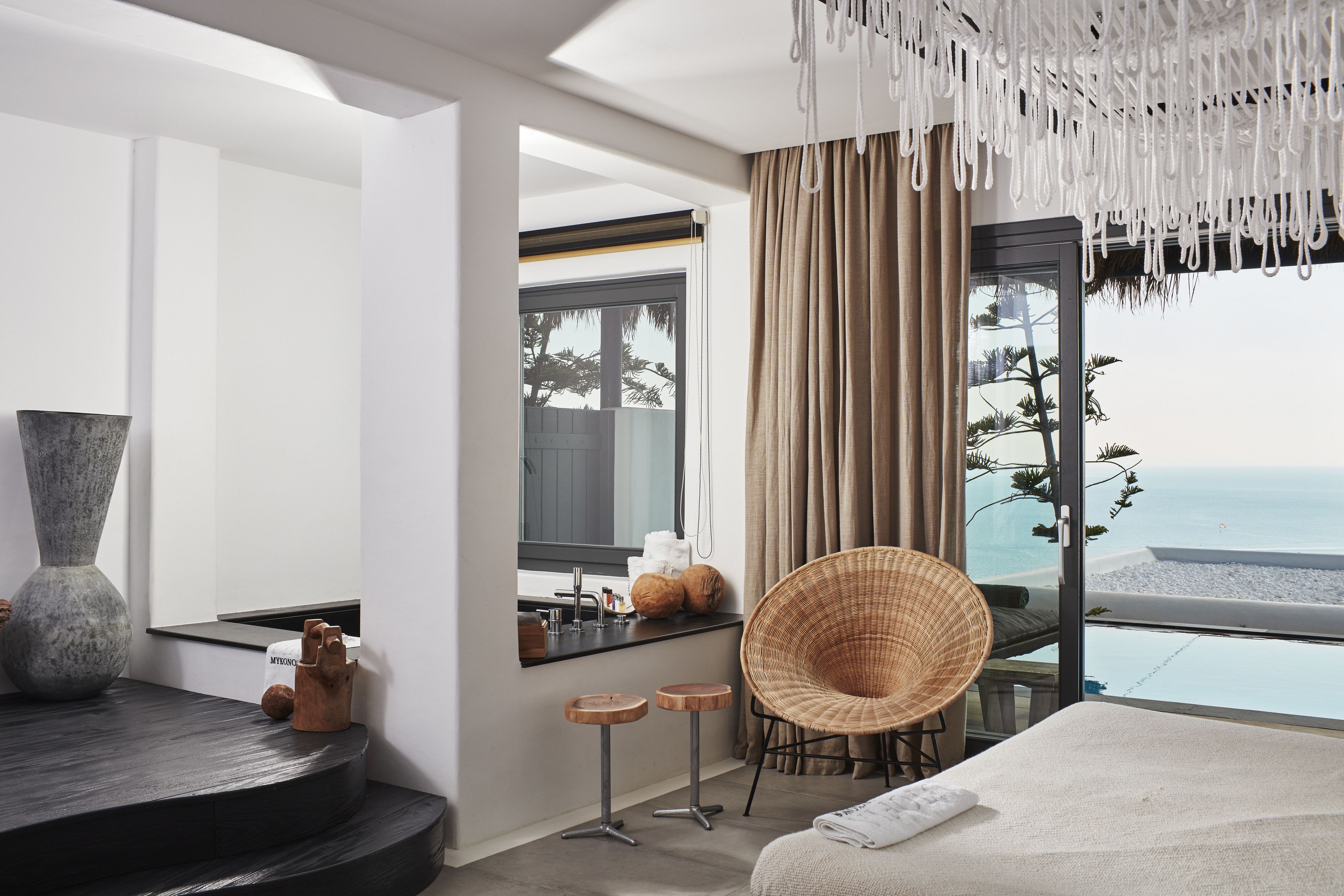A modern hotel room with a large window offering a view of the beach, a comfortable chair, a vanity with a mirror, and a large bed.